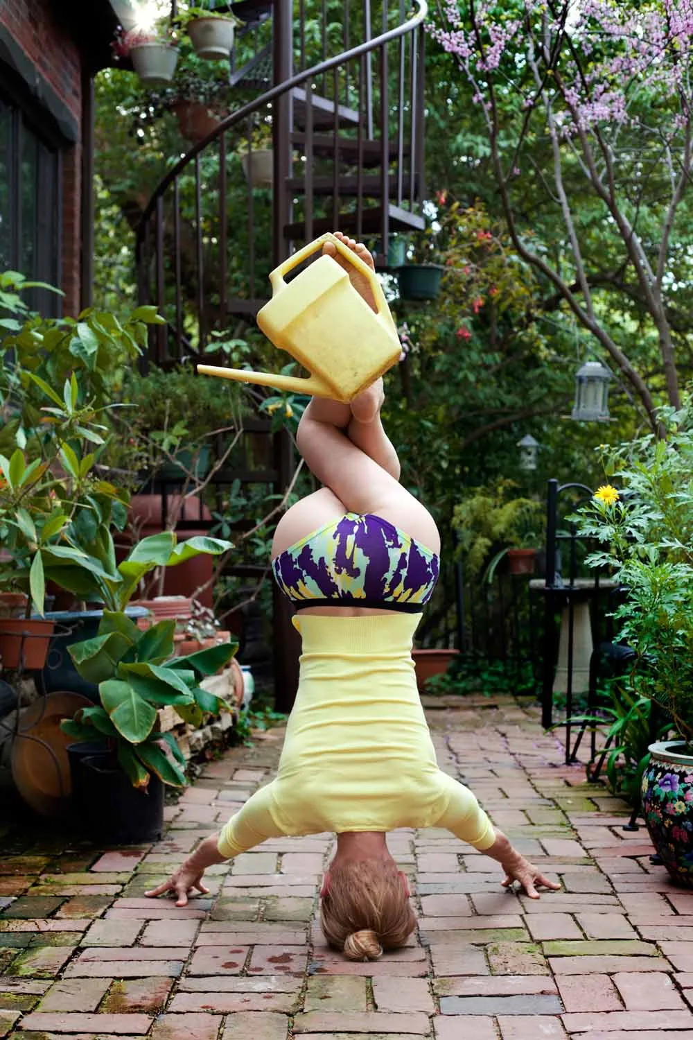 Michelle in a headstand on a brick garden path, balancing a yellow watering can on the feet